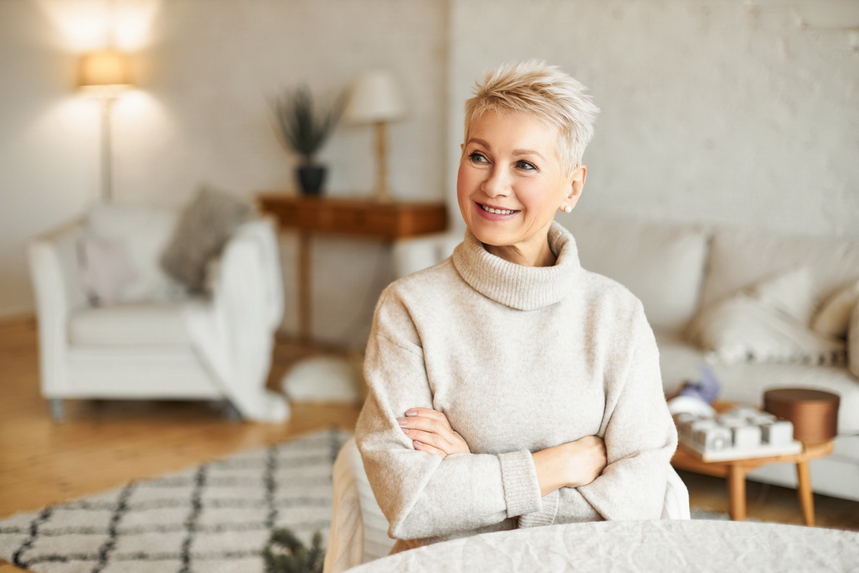 Portrait of gorgeous mature European female with short hairstyle relaxing at home sitting at table in living room crossing arms on chest trying to warm up in cozy cashmere turtleneck sweater, smiling